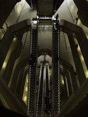 A set of escalators viewed from above, located in a spacious, modern indoor environment with multiple levels. The architectural design features smooth lines and a combination of metallic and dark shades. People are seen travelling up and down on the escalators, giving a sense of movement and scale.