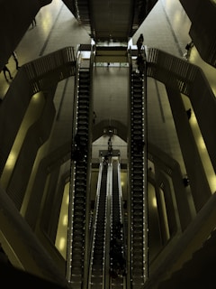 A set of escalators viewed from above, located in a spacious, modern indoor environment with multiple levels. The architectural design features smooth lines and a combination of metallic and dark shades. People are seen travelling up and down on the escalators, giving a sense of movement and scale.
