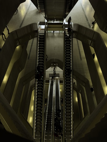 A set of escalators viewed from above, located in a spacious, modern indoor environment with multiple levels. The architectural design features smooth lines and a combination of metallic and dark shades. People are seen travelling up and down on the escalators, giving a sense of movement and scale.