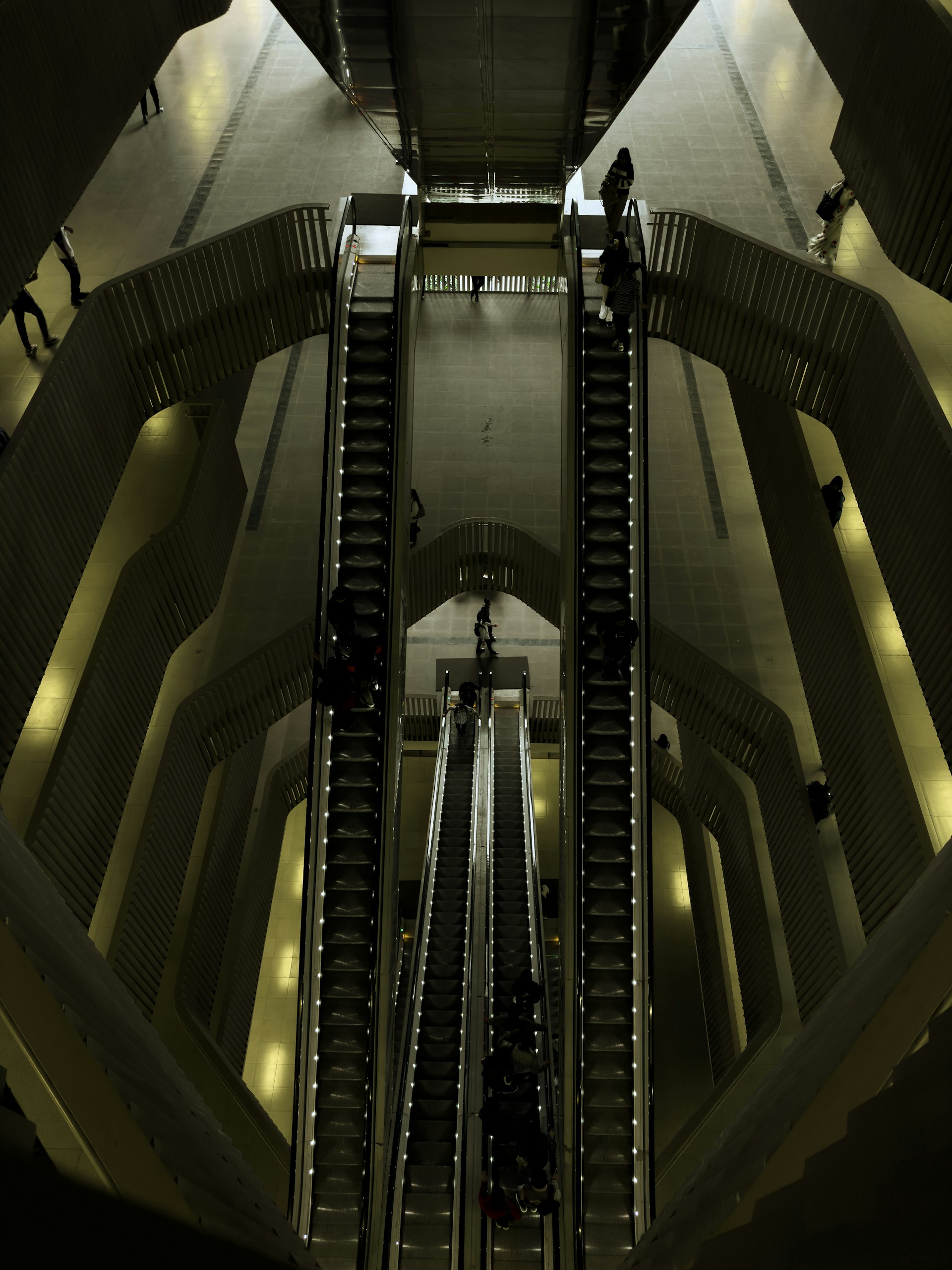 A set of escalators viewed from above, located in a spacious, modern indoor environment with multiple levels. The architectural design features smooth lines and a combination of metallic and dark shades. People are seen travelling up and down on the escalators, giving a sense of movement and scale.