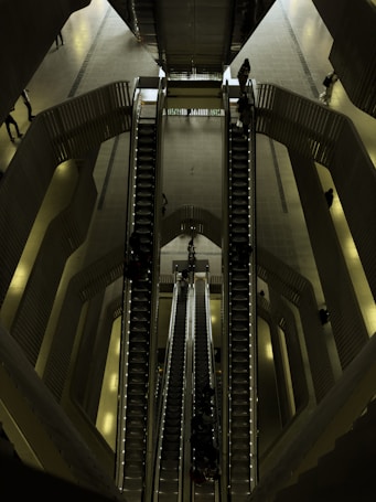 A set of escalators viewed from above, located in a spacious, modern indoor environment with multiple levels. The architectural design features smooth lines and a combination of metallic and dark shades. People are seen travelling up and down on the escalators, giving a sense of movement and scale.