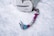 A pink and teal ice axe lies on a snowy surface next to a white backpack. The axe has a curved handle with a sturdy grip, designed for climbing. The backpack appears worn and is lightly dusted with snow.