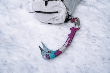 A pink and teal ice axe lies on a snowy surface next to a white backpack. The axe has a curved handle with a sturdy grip, designed for climbing. The backpack appears worn and is lightly dusted with snow.