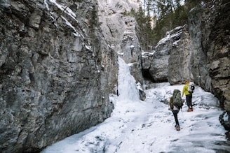 Two hikers equipped with backpacks and crampons navigate a snowy, icy path through a narrow canyon surrounded by towering rock walls. Evergreen trees are visible at the top of the cliffs, and the icy terrain indicates a cold, rugged environment.