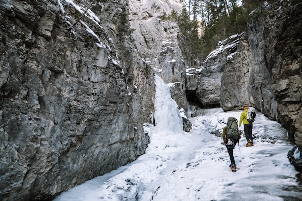 Two hikers equipped with backpacks and crampons navigate a snowy, icy path through a narrow canyon surrounded by towering rock walls. Evergreen trees are visible at the top of the cliffs, and the icy terrain indicates a cold, rugged environment.