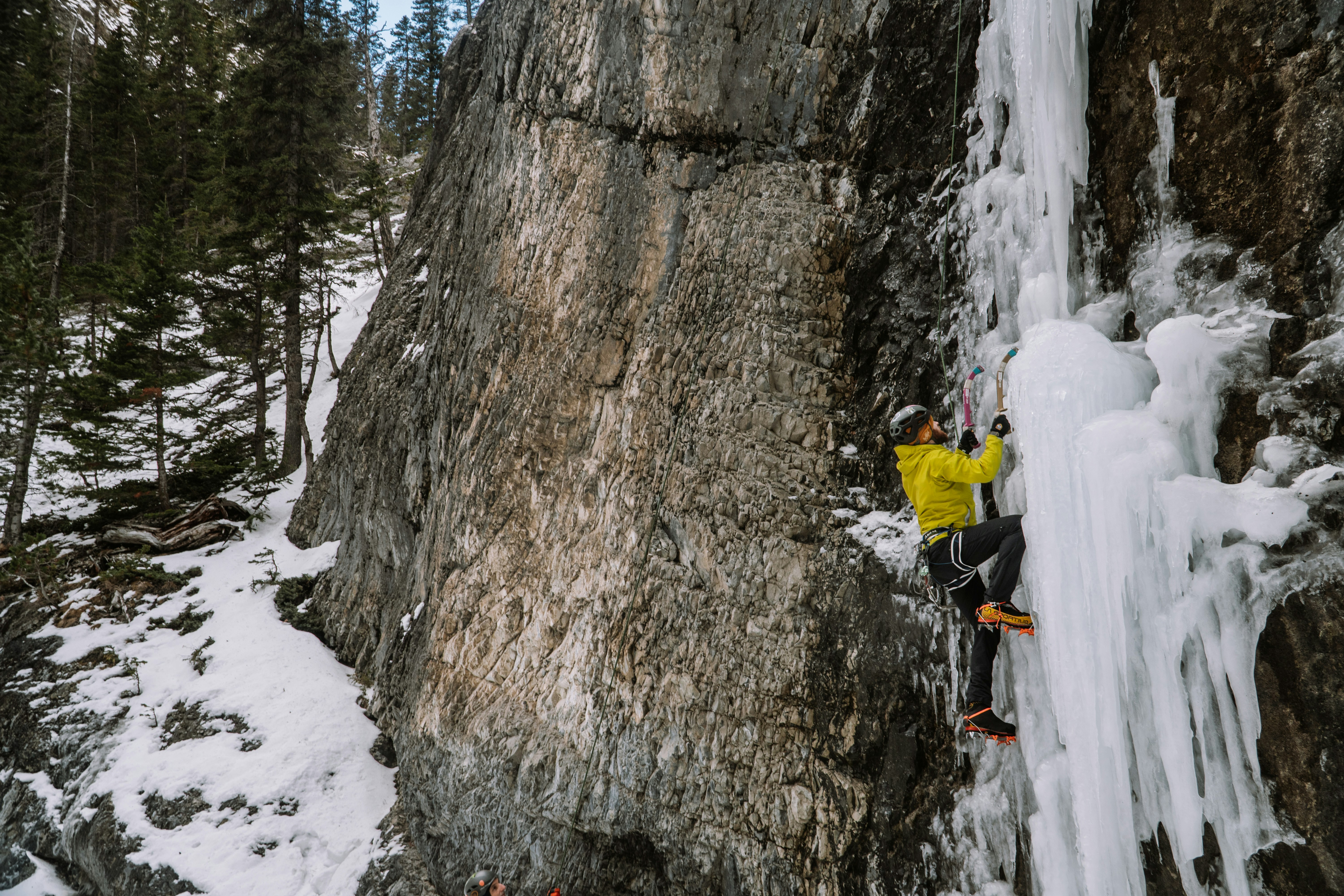 An ice climber tests a placement high on a vertical wall of blue ice, ice tool raised for the next hold
