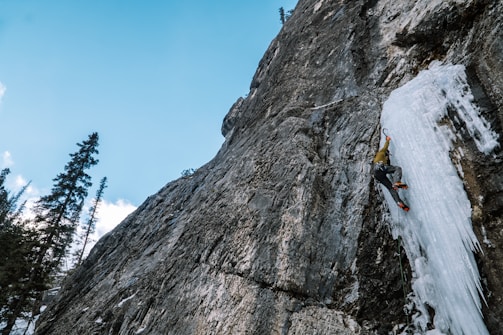 A climber is ascending a steep, icy rock face using ice axes and crampons. The sky is clear with a few clouds, and tall pine trees are visible on the left side.