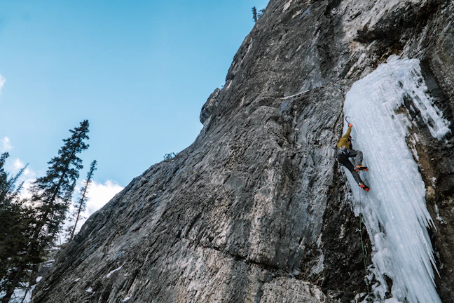 A climber gripping an ice axe, poised on a steep icy slope under a clear blue sky.