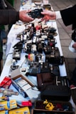 A variety of vintage cameras and some film-related items are displayed on a white tablecloth at a market or street sale. Two people are exchanging one of the cameras, with their hands reaching out towards each other. The table is crowded with different types of cameras and camera equipment, along with some boxes of photo paper and miscellaneous items.