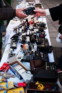 A variety of vintage cameras and some film-related items are displayed on a white tablecloth at a market or street sale. Two people are exchanging one of the cameras, with their hands reaching out towards each other. The table is crowded with different types of cameras and camera equipment, along with some boxes of photo paper and miscellaneous items.
