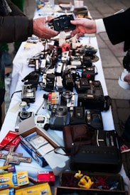 A variety of vintage cameras and some film-related items are displayed on a white tablecloth at a market or street sale. Two people are exchanging one of the cameras, with their hands reaching out towards each other. The table is crowded with different types of cameras and camera equipment, along with some boxes of photo paper and miscellaneous items.