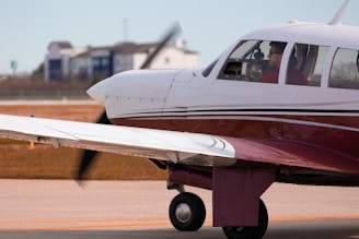 A small propeller airplane is on a runway, with its engine running. The aircraft is primarily white with a maroon stripe and has a single propeller on the front. A person is visible inside the cockpit, wearing sunglasses and a red shirt. The background shows some buildings and a grassy area.