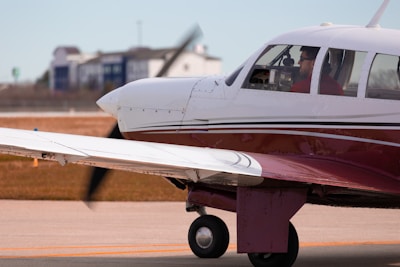 A small propeller airplane is on a runway, with its engine running. The aircraft is primarily white with a maroon stripe and has a single propeller on the front. A person is visible inside the cockpit, wearing sunglasses and a red shirt. The background shows some buildings and a grassy area.
