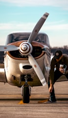 A small propeller airplane is parked on the tarmac with a person working on the ground near the front wheel. The aircraft has a distinctive spiral pattern on the propeller and is painted in white and brown. The background shows a clear blue sky and distant trees.