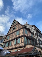 A traditional European timber-framed house with beige and brown wooden beams, featuring light turquoise window shutters. The building has a steep triangular roof and is set against a partly cloudy blue sky.