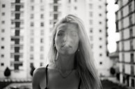 A black and white portrait photo featuring a woman with long blonde hair standing in front of a blurred background of apartment buildings. The focus is on her face, with a neutral or contemplative expression.