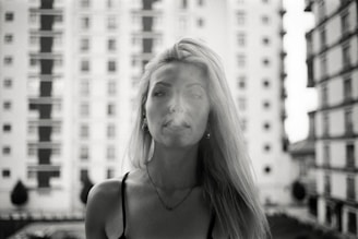 A black and white portrait photo featuring a woman with long blonde hair standing in front of a blurred background of apartment buildings. The focus is on her face, with a neutral or contemplative expression.