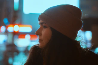 Close-up of a hand adjusting a neon-striped beanie with a blurred urban nightscape in the background.