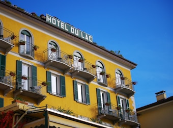 A vibrant yellow building with multiple balconies adorned with potted plants and flowers. The windows are framed with dark green shutters. The top of the building displays a sign labeled 'Hotel Du Lac.' The sky is clear and blue, adding a bright and cheerful ambiance.