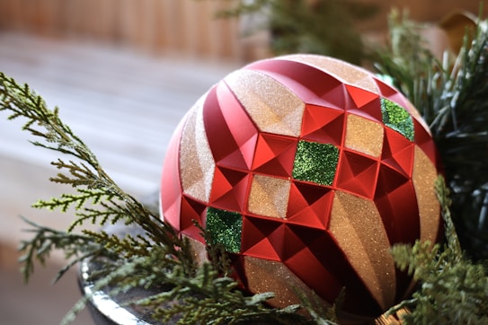 A decorative Christmas ornament featuring a geometric pattern with red and gold sections, embellished with green glitter squares. The ornament is nestled among green pine branches, adding a festive touch.