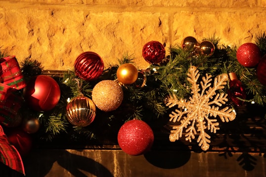 A festive arrangement featuring various Christmas ornaments including red, gold, and glittery spheres, a large gold snowflake, and a red-green bow. These decorations are nestled in a garland of green pine needles, set against a textured stone wall, creating a warm, holiday atmosphere.