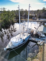 A large sailboat docked at a small pier in a serene lake area surrounded by dense green trees. A truck with a crane is parked nearby, possibly for maintenance or construction work. The water is calm, reflecting the trees and sky.
