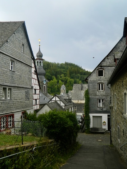 A quaint village street is lined with traditional European architecture, featuring slate-roofed buildings and timber framing. A church steeple rises in the background against a backdrop of lush green hills and trees, conveying a picturesque and serene rural setting.