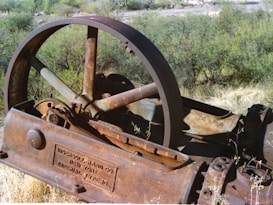 An old, rusted piece of industrial machinery with an engraved plaque showing Ingersoll-Rand Co., New York, Imperial Type 10. The machinery appears to be abandoned and situated in a natural environment with dry grass and various green shrubs.