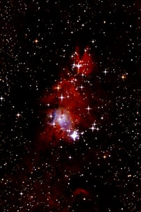 A celestial scene filled with numerous bright stars scattered across a dark expanse. At the center, a rich reddish nebula emits vibrant colors, partially illuminated by nearby stars. wisps of nebular clouds create a sense of depth and majesty in the cosmic environment.