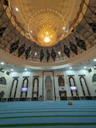 An ornate mosque interior featuring a large, intricate chandelier dominating the ceiling space. The architecture showcases arched windows and decorative white walls with geometric patterns. The prayer area is covered with blue carpets having green stripes. Two digital screens on the walls display information.