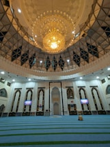 An ornate mosque interior featuring a large, intricate chandelier dominating the ceiling space. The architecture showcases arched windows and decorative white walls with geometric patterns. The prayer area is covered with blue carpets having green stripes. Two digital screens on the walls display information.