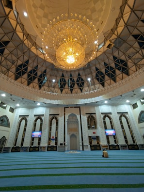 An ornate mosque interior featuring a large, intricate chandelier dominating the ceiling space. The architecture showcases arched windows and decorative white walls with geometric patterns. The prayer area is covered with blue carpets having green stripes. Two digital screens on the walls display information.