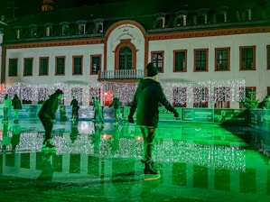 Ice skaters glide on a rink in front of a building adorned with festive lights. The scene is illuminated with a green hue, adding a magical atmosphere. The building has numerous windows and an ornate facade, creating a charming backdrop.