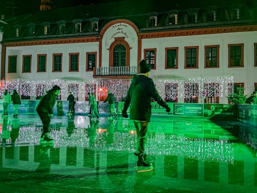 Ice skaters glide on a rink in front of a building adorned with festive lights. The scene is illuminated with a green hue, adding a magical atmosphere. The building has numerous windows and an ornate facade, creating a charming backdrop.