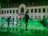 Ice skaters glide on a rink in front of a building adorned with festive lights. The scene is illuminated with a green hue, adding a magical atmosphere. The building has numerous windows and an ornate facade, creating a charming backdrop.