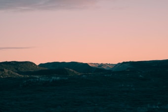 A landscape with dark, rugged hills in the foreground and a smooth, pastel-colored sky transitioning from orange to pale pink. The scene conveys a serene and tranquil atmosphere, capturing the subtle beauty of a sunset.