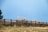 A wooden fence stretches diagonally across a grassy field. Tall grasses and a few green shrubs can be seen behind the fence, with a bright blue sky above and snow gently falling.