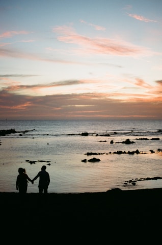 A soft rose-colored backdrop with a silhouette of a woman gently holding a man's hand on a beach at sunset.