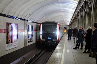 A metro train is arriving at an underground station, where several passengers are waiting on the platform. The station features a vaulted ceiling and advertising posters on the walls. The train has a modern design with headlights on, illuminating the dimly lit platform.