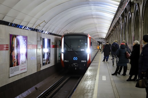 A metro train is arriving at an underground station, where several passengers are waiting on the platform. The station features a vaulted ceiling and advertising posters on the walls. The train has a modern design with headlights on, illuminating the dimly lit platform.