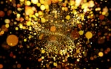 A multitude of shimmering, glowing particles in various sizes and shades of golden yellow, orange, and brown are scattered across a dark background. The particles create a vibrant, dynamic scene, resembling a burst of light or confetti.