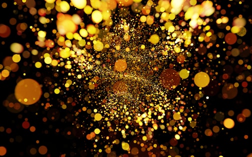 A multitude of shimmering, glowing particles in various sizes and shades of golden yellow, orange, and brown are scattered across a dark background. The particles create a vibrant, dynamic scene, resembling a burst of light or confetti.