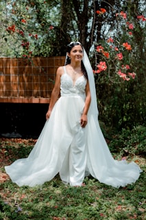 A bride stands outdoors wearing a flowing white wedding dress and veil, surrounded by lush greenery and vibrant red flowers. Sunlight filters through the trees, creating a serene and romantic atmosphere.