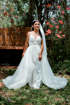 A sunlit portrait of a bride in a flowing ivory dress standing amid soft green foliage, embodying timeless elegance and serenity.