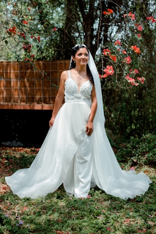 A bride stands outdoors wearing a flowing white wedding dress and veil, surrounded by lush greenery and vibrant red flowers. Sunlight filters through the trees, creating a serene and romantic atmosphere.