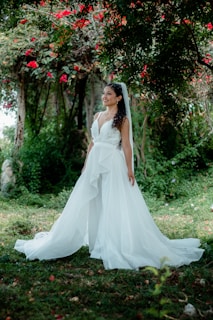 A bride in a flowing white wedding dress stands in a lush garden setting, surrounded by vibrant greenery and bright red flowers. Her curly hair is styled elegantly with a veil, and she has an expression of joy and serenity.