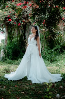 A bride in a flowing white wedding dress stands in a lush garden setting, surrounded by vibrant greenery and bright red flowers. Her curly hair is styled elegantly with a veil, and she has an expression of joy and serenity.