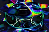 A large transparent lens reflects and refracts bright, rainbow-colored patterns on a dark background. The vibrant spectrum of colors creates a dynamic and mesmerizing visual effect.