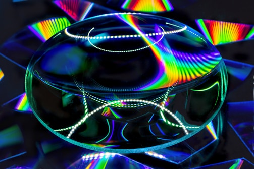 A large transparent lens reflects and refracts bright, rainbow-colored patterns on a dark background. The vibrant spectrum of colors creates a dynamic and mesmerizing visual effect.