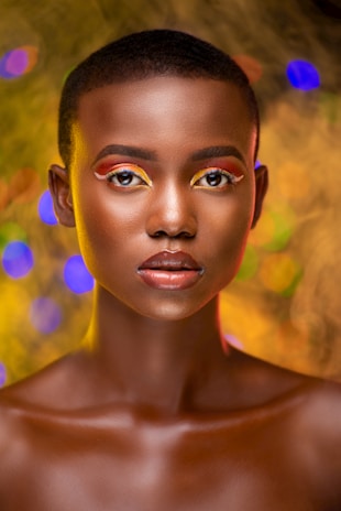 A model with a shaved head and glowing skin is centered in the image, wearing striking makeup with vibrant colors around the eyes, including red and yellow eyelids with precise eyeliner. The background features soft, colorful bokeh lights in shades of blue, orange, and green, creating an artistic and dreamy effect.
